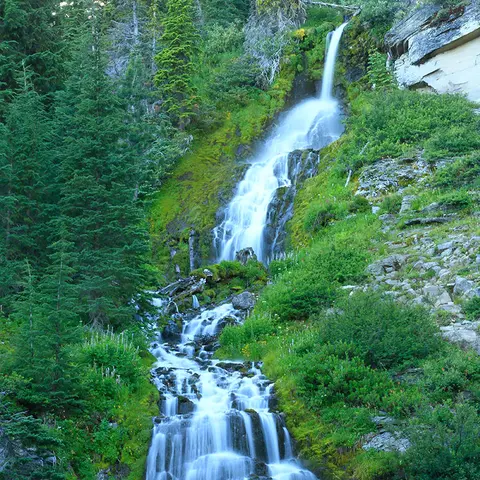 A waterfall flows down a rocky, green hillside surrounded by trees.