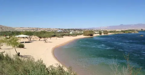A sandy beach next to a river with clear blue water and distant hills.