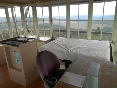 An interior view of a fire lookout tower with a bed, desk, and large windows overlooking a mountainous landscape.