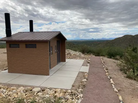 An outdoor restroom building sits along a desert path under a cloudy sky.