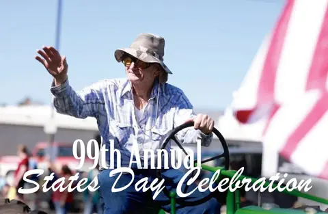 An older man wearing a hat and sunglasses waves while driving a tractor in a parade.
