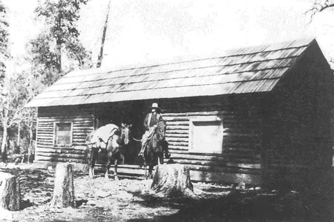 Black and white historic photo showing a person on a horse outside a log cabin.