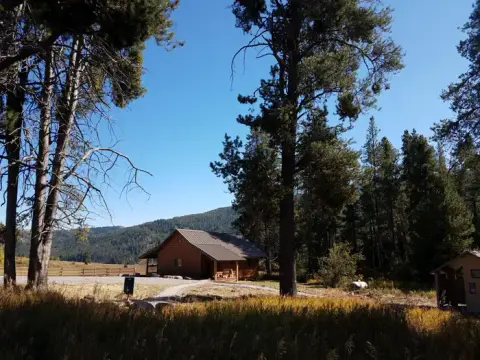 A cabin nestled among tall pine trees and mountains under a clear blue sky.