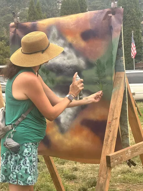 A person paints on a canvas outdoors using a spray can and a plant as a stencil.