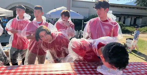 A group of young men wearing plastic ponchos participate in an outdoor pie-eating contest.