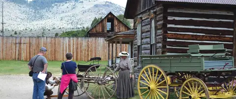 People dressed in period costume walk near historic log buildings and horse-drawn wagons.