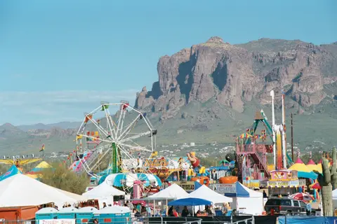 A colorful carnival is set up with rides and tents in front of a large mountain.