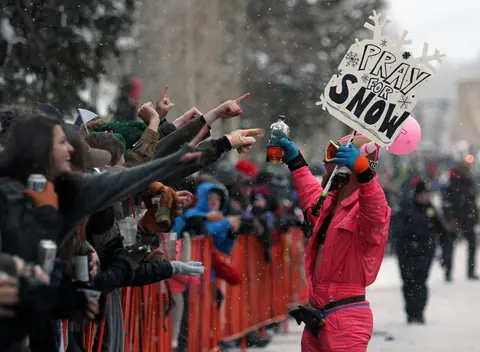 A person in a pink ski suit holds a sign and a bottle before a crowd in the snow.