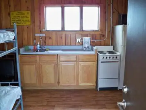 An interior view of a cabin kitchen with wooden paneling, a sink, stove, and bunk beds.