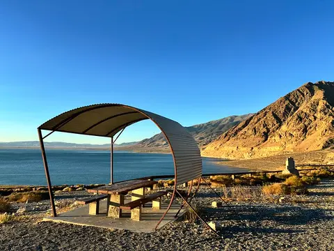 A shaded picnic table overlooks a large body of water and mountains.