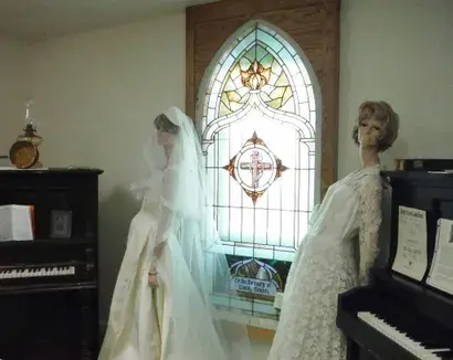 Two mannequins in wedding dresses stand in an interior room with a stained glass window.