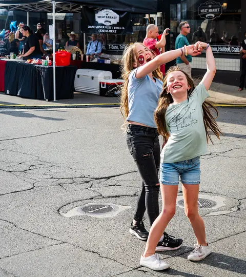 Two girls dancing outdoors on a street with tents and people in the background.