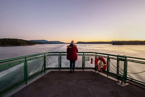 A person stands at the stern of a ferry looking out at the water and distant shores.