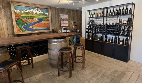 A rustic wine tasting room with a wooden table, barrels, stools, and wine displays.