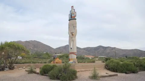 A large statue stands upright in an outdoor desert landscape with mountains in the background.