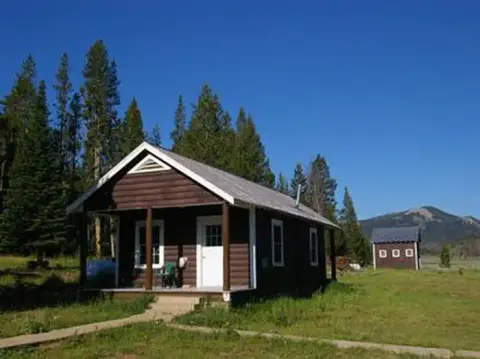 A small brown cabin with a white door and covered porch sits in a grassy clearing surrounded by trees under a clear blue sky.