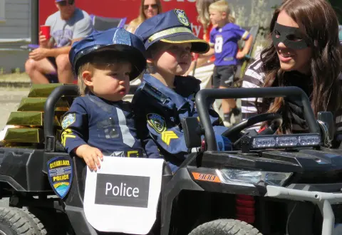 Two children dressed as police officers sit in a toy vehicle with an adult wearing a mask.