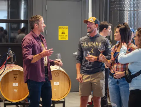 People hold wine glasses and talk inside a building with barrels.