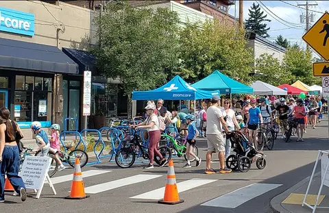 A photograph showing a crowd of people walking and biking along a street with tents and shops.