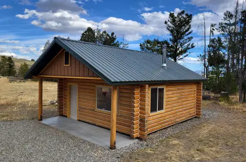 A log cabin with a metal roof and a porch stands outdoors under a cloudy sky.