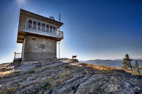 A fire lookout tower stands on a rocky mountain peak with a dog looking out over the landscape.
