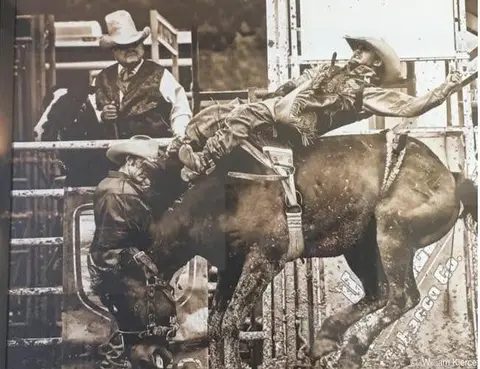 A black and white photograph shows a cowboy riding a bucking horse at a rodeo.
