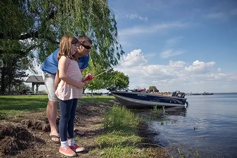 An adult and a young girl fish together by the water's edge.