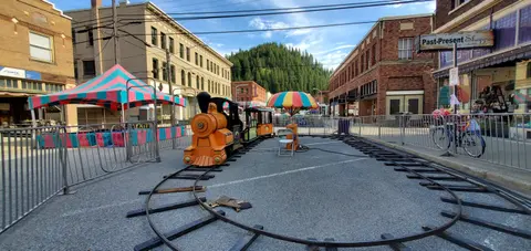 A small toy train sits on tracks set up on a street between brick buildings.
