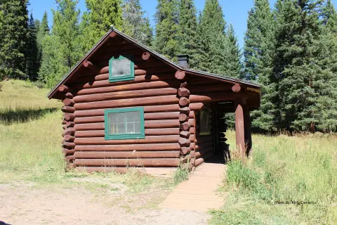 A log cabin sits in a clearing surrounded by tall evergreen trees.