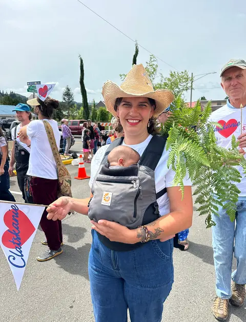 A woman wearing a cowboy hat and holding a baby in a carrier poses outside during a parade or street event.