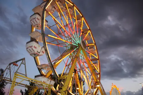A brightly lit Ferris wheel is seen against a dark, cloudy sky at dusk.