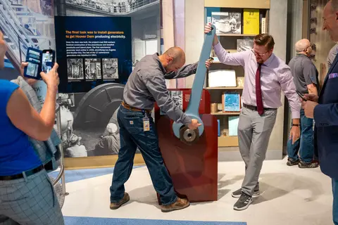 Two men pose while pretending to turn a large wrench exhibit inside a museum.