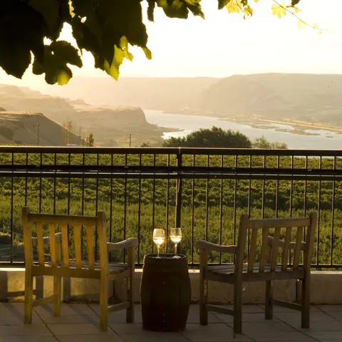 Two wine glasses on a small barrel between two chairs on a balcony overlooking a landscape with water.