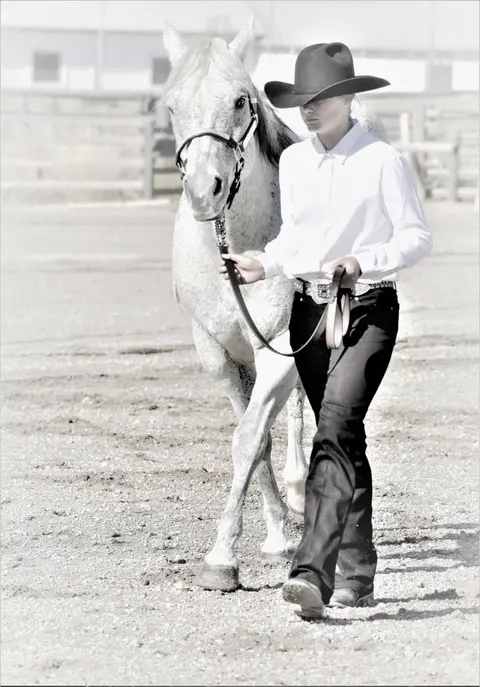 A person in a cowboy hat and white shirt leads a horse outdoors.