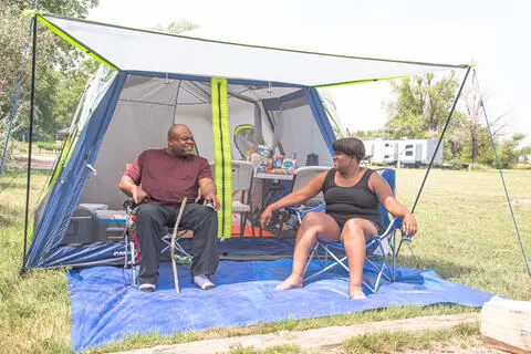Two people sit outside a tent set up on a lawn.