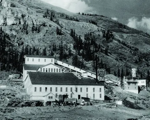A historic black and white photo shows a large industrial mill nestled against a rocky mountain slope with several old cars parked in front.