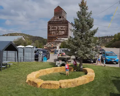 An old wooden grain elevator stands near a grassy area with people and parked vehicles.