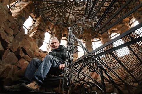 A man sits on a spiral metal staircase inside a stone tower.