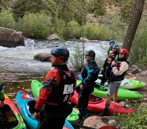 Kayakers in helmets and wetsuits stand by a rushing river with their kayaks on the shore.