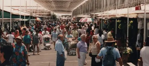 A crowded outdoor market scene with people walking between vendor stalls.