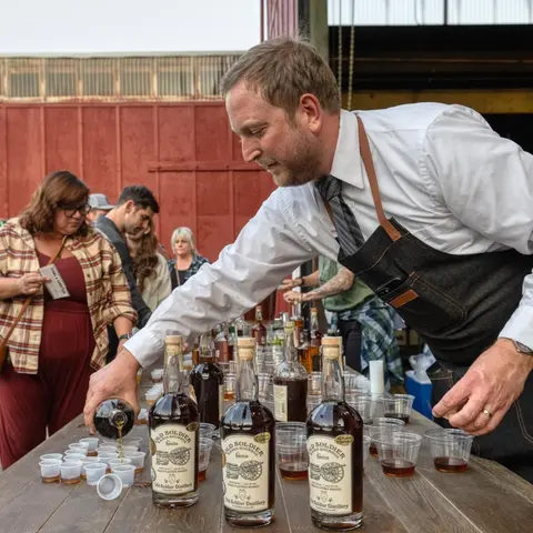 A man in an apron pours drinks from a bottle into small cups on a table.