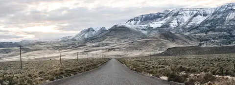 A straight asphalt road stretches toward snow-dusted mountains under a cloudy sky.