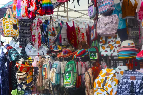 A market stall displays an assortment of colorful clothing, backpacks, and hats.