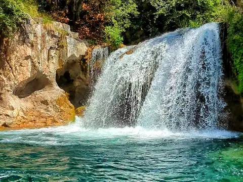 A waterfall flows into a clear pool surrounded by rocky cliffs and green trees.