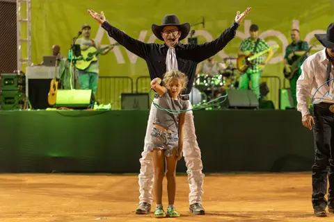 A cowboy stands behind a young girl holding a rope on a stage with a band in the background.