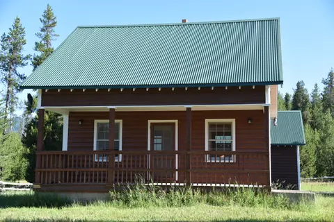 A brown cabin with a green corrugated roof and front porch stands among trees and grass.
