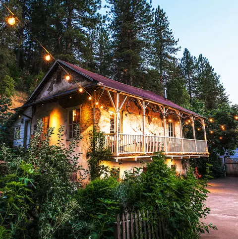 A rustic two-story building surrounded by trees is illuminated by string lights at dusk.