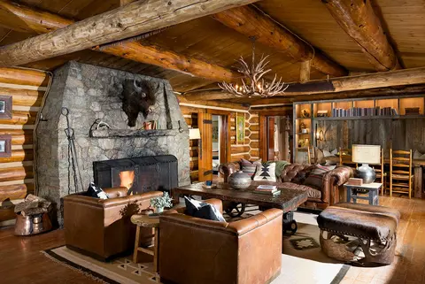 An interior view of a rustic log cabin living room with a stone fireplace and leather furniture.