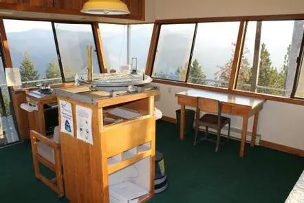 The interior of a fire lookout tower with panoramic windows overlooking forest and mountains.
