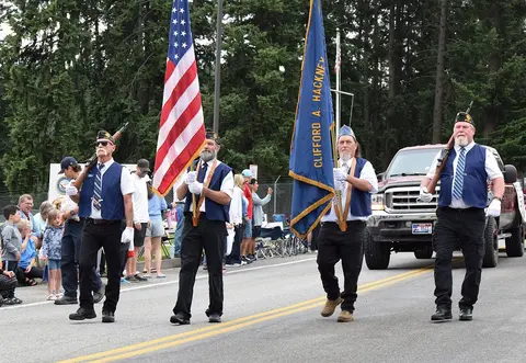 People march in a parade carrying flags and rifles on a street.